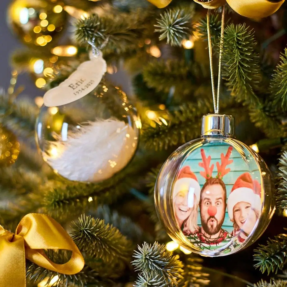 Close-up of a decorated Christmas tree featuring a clear bauble with a festive design and gold ribbon accents, alongside another ornament containing a white feather.